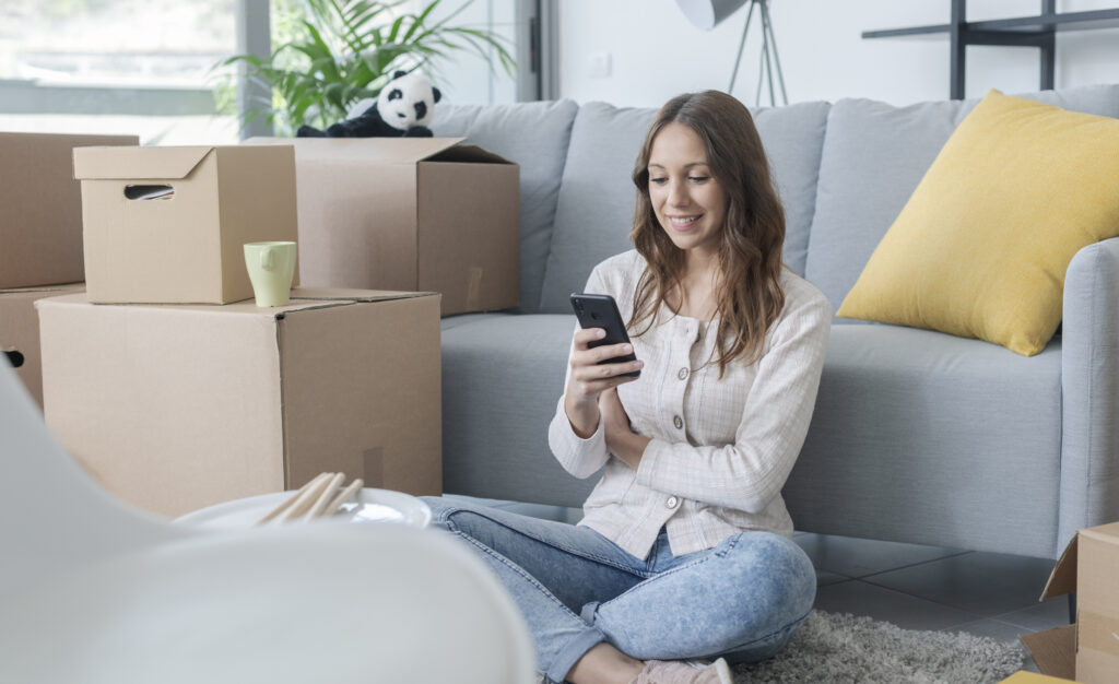 Happy woman in her new apartment, sitting on the floor and chatting on her smartphone