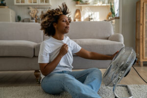 A woman sits on the floor using a fan to cool off from the summer heat. 
