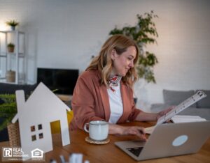 A property manager sits at a table with a laptop and a small house model, analyzing maintenance data.