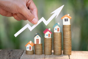 A hand holding an arrow cut-out above tiny houses on coin stacks, illustrating the concept of investing in real estate.