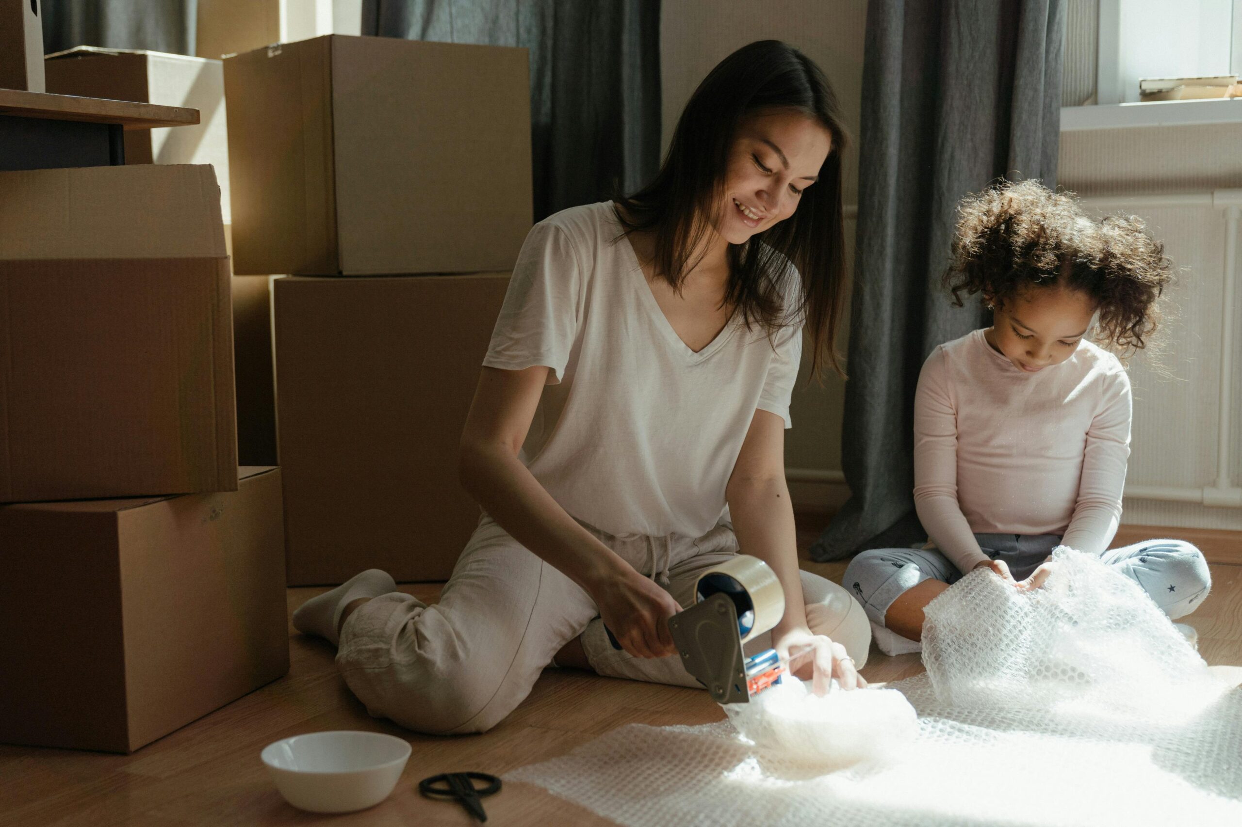 A mother and child sitting on the floor of their home surrounded by moving boxes.
