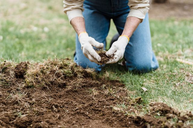 a-person-in-jeans-and-gloves-kneeling-on-grass-tending-to-soil-in-a-garden-setting