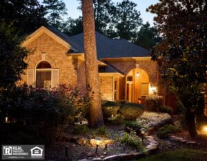 Exterior of a lovely stone and brick house surrounded by trees and a flourishing garden at night.
