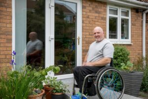 A man in a wheelchair outside his home on a wheelchair ramp, sitting on a cozy patio.