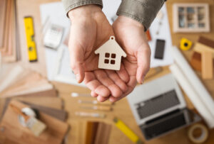 Real estate agent holding a small house, desktop with tools, wood swatches and computer on background, top view. 