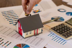 Hand placing coin on house model on table with financial documents.
