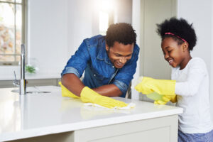 A father and his daughter smiling and wearing cleaning gloves, happily cleaning the kitchen together.
