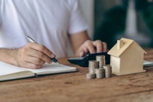 A man drafting a maintenance budget in a notebook with a pen, next to a house model on the table.