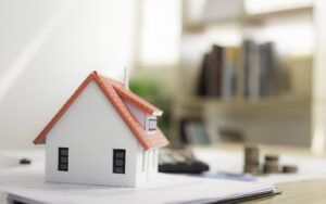 A desk with a house model atop some papers, and a stack of coins in the background, highlighting property pricing.