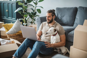 Man sitting on floor, with his dog and laptop,amongst moving boxes.
