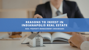 A miniature model house sits on a book, symbolizing real estate investment. A suited person reviews documents in the background. Text: "Reasons to Invest in Indianapolis Real Estate."
