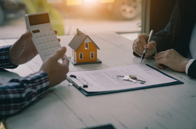 Two individuals at a table discuss real estate, featuring a model house, a contract with a pen and keys, and a calculator.