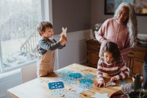 Senior woman works on a DIY winter craft with her two young grandchildren, at home in the dining room.