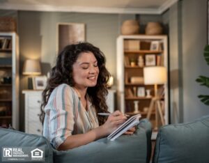 Young woman with long brown hair sits comfortably on her sofa at home, smiling as she writes in a notebook.