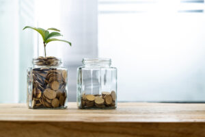 Two jars filled with coins, one with a green plant, on a wooden table.