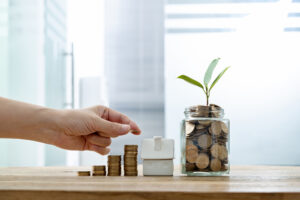 A hand placing coins on a table with a house and a plant in a 
jar.
