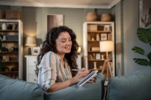 Young woman with long brown hair sits comfortably on her sofa at home, smiling as she writes in a notebook.