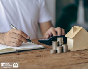 A man drafting a maintenance budget in a notebook with a pen, next to a house model on the table.