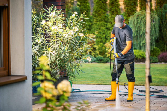 Pressure Washing a Patio in a Residential Backyard During a Sunny Afternoon With Well-Maintained Plants and Greenery Surrounding the Area A person wearing yellow rubber boots and a gray cap uses a pressure washer to clean a patio in a residential backyard. The sun shines brightly on the scene, highlighting the manicured lawn and vibrant plants around.