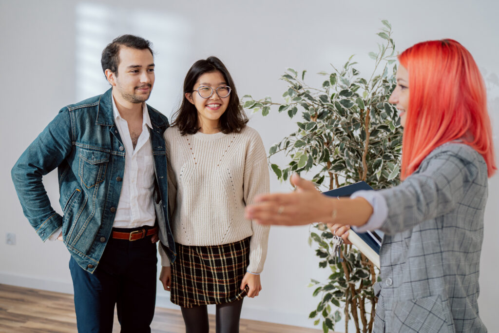 Happy couple stand in empty room while inspecting apartment for rent with real estate agent woman and man look at place indicated by pink-haired girl touring rooms pointing out equipment appliances