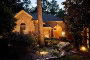Exterior of a lovely stone and brick house surrounded by trees and a flourishing garden at night.
