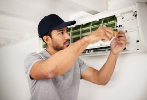 A man is working on an air conditioner, focused on using a screwdriver.
