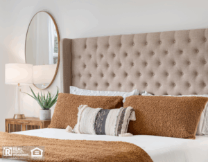 A bedroom detail shot with a cushioned headboard, a gold mirror on the wall, and a plant and lamp on the nightstand.