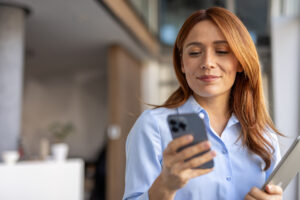 A woman in a blue shirt holds a tablet while checking her phone, looking focused and engaged.