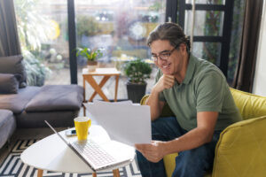 A man sitting in a yellow chair reviewing papers and working on a laptop.