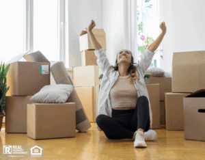 Young woman sitting on floor in new apartment with boxes and raising arms in joy.