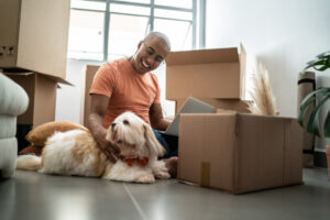 A man and his dog are seated on the floor, surrounded by boxes in their new home.
