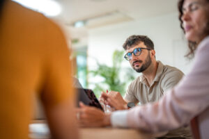 A man wearing glasses is seated at a table with two others, engaged in a friendly discussion. 