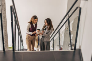 Two businesswomen on stairs in an office building, discussing plans for a rental property. 