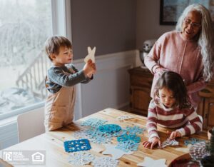 Senior woman works on a DIY winter craft with her two young grandchildren, at home in the dining room.