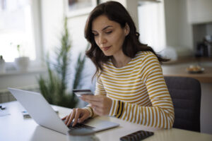 A woman sitting at a table, using a laptop while holding a credit card in her hand. 