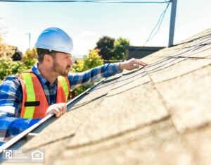 Man with hard hat standing on ladder inspecting house roof.