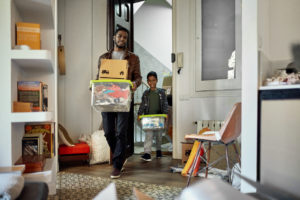 Afro-Caribbean single father in late 20s and 7 year old son smiling as they carry cardboard boxes and plastic containers into new home.