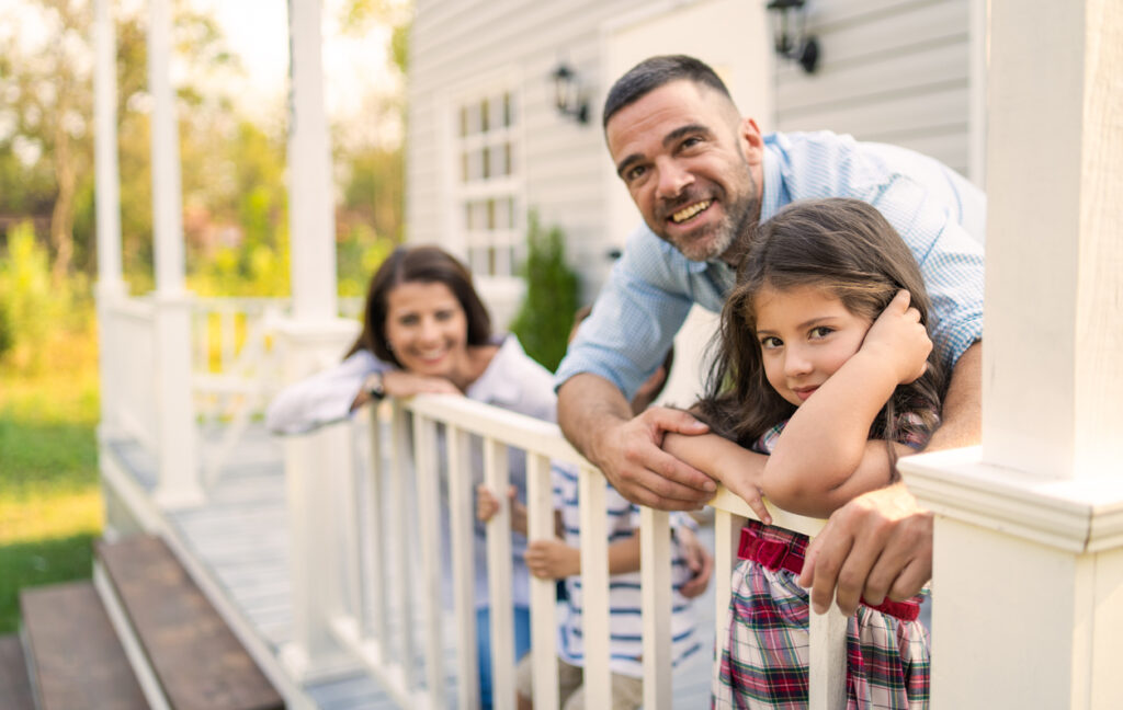 A family of four standing on the front porch and leaning on the railings while smiling at the camera outside on a bright day