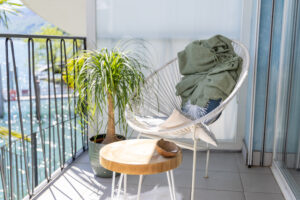 Cozy balcony featuring a modern chair with a green throw, a potted plant, and a small wooden table in bright sunlight. 