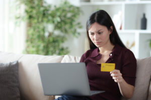 A woman seated on a couch, anxiously holding a credit card, reflecting the tension of a potential rental scam situation.

